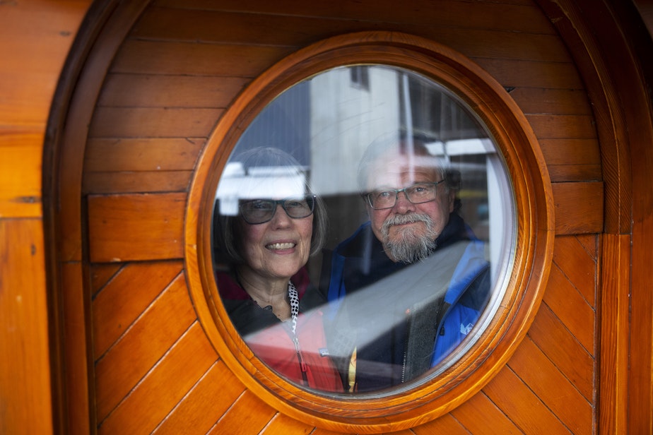 caption: Janet Yoder and Robert Rudine are portrayed looking through a port hole window on their houseboat, the self declared sovereign nation of Tui Tui, on Tuesday, March 26, 2024, in Seattle. 