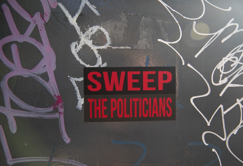 caption: A sticker that reads 'Sweep The Politicians' is shown near Denny Park as unhoused people are swept from the area by the city of Seattle, on Wednesday, March 3, 2021.