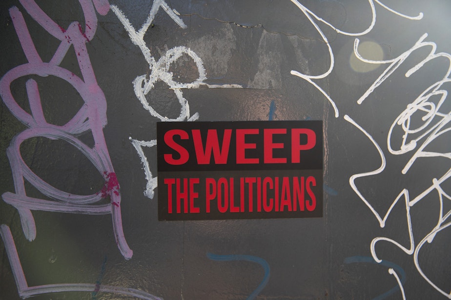 caption: A sticker that reads 'Sweep The Politicians' is shown near Denny Park as unhoused people are swept from the area by the city of Seattle, on Wednesday, March 3, 2021. 