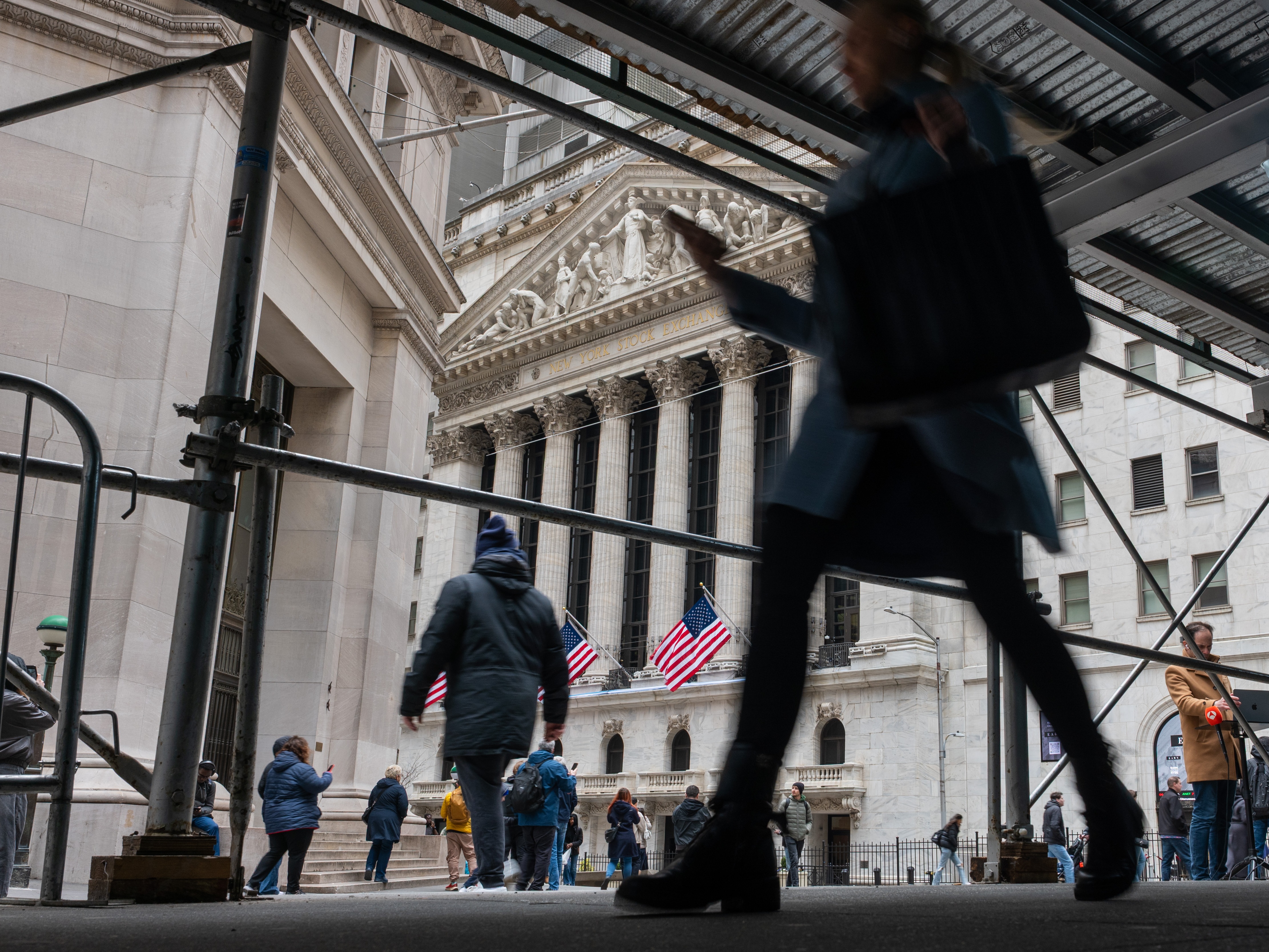 caption: People walk by the New York Stock Exchange on Wall Street. Trades of tariff refunds have ballooned since the Supreme Court struck down many of President Trump's tariffs.