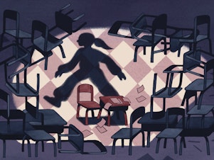 The shadow of a student walks away from an empty desk surrounded by more empty chairs and desks in a school classroom.