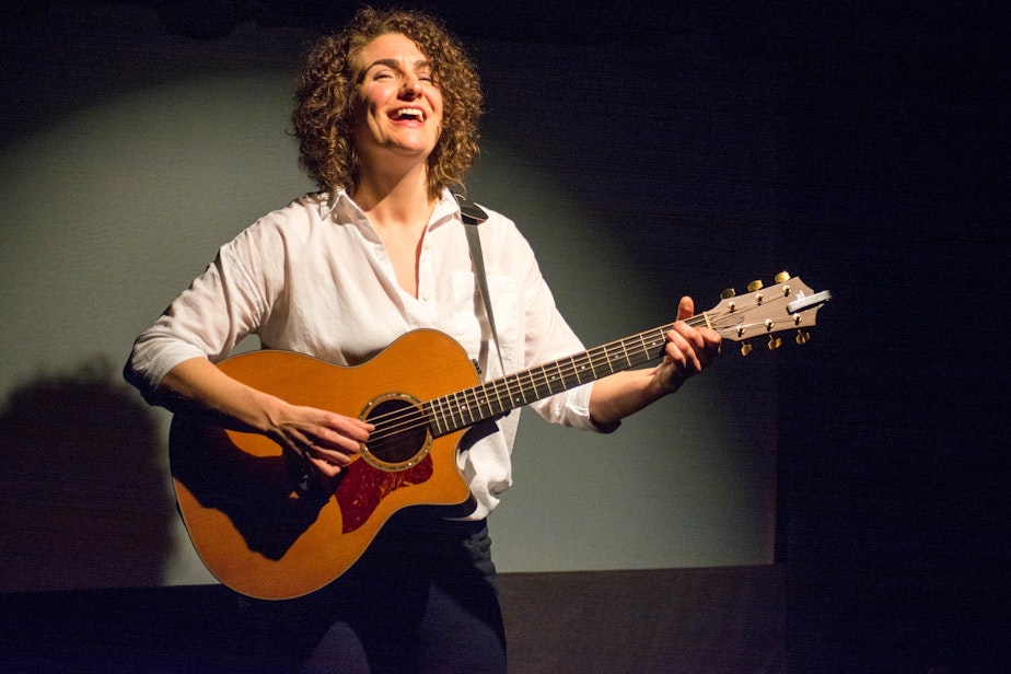 caption: Bridget Quigg plays guitar on stage during her one-woman show 'Techlandia.'