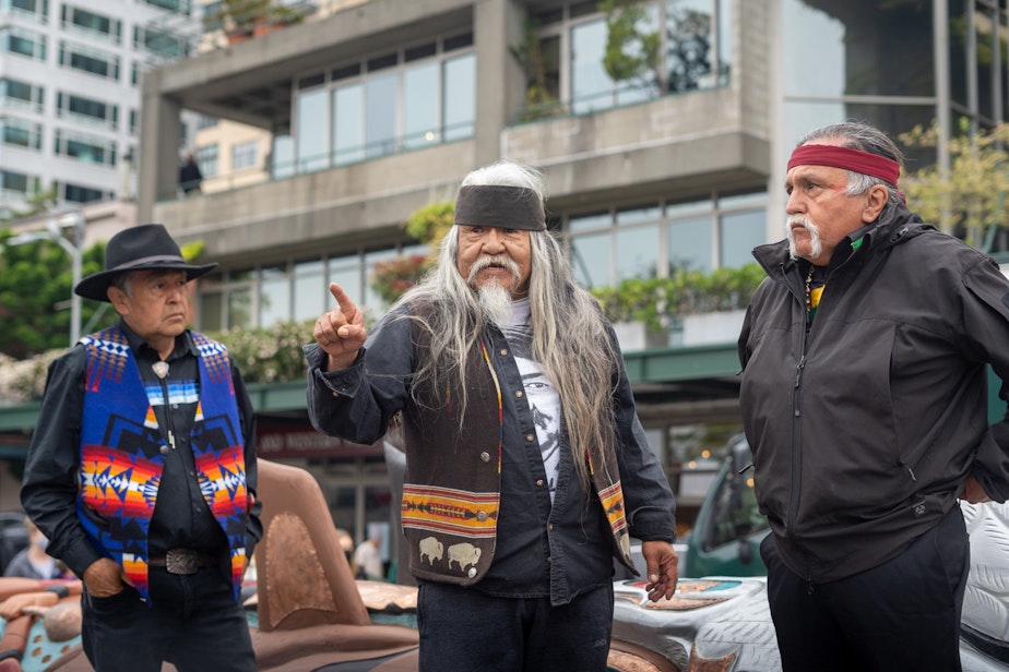 caption: Master carvers from left, Douglas (Sit-ki-kadem) James, Rick Williams, and Jewell Praying Wolf James stand in front of the 24-foot totem pole while addressing the crowd on Saturday, May 22, 2021, outside of Seattle’s Pike Place Market.