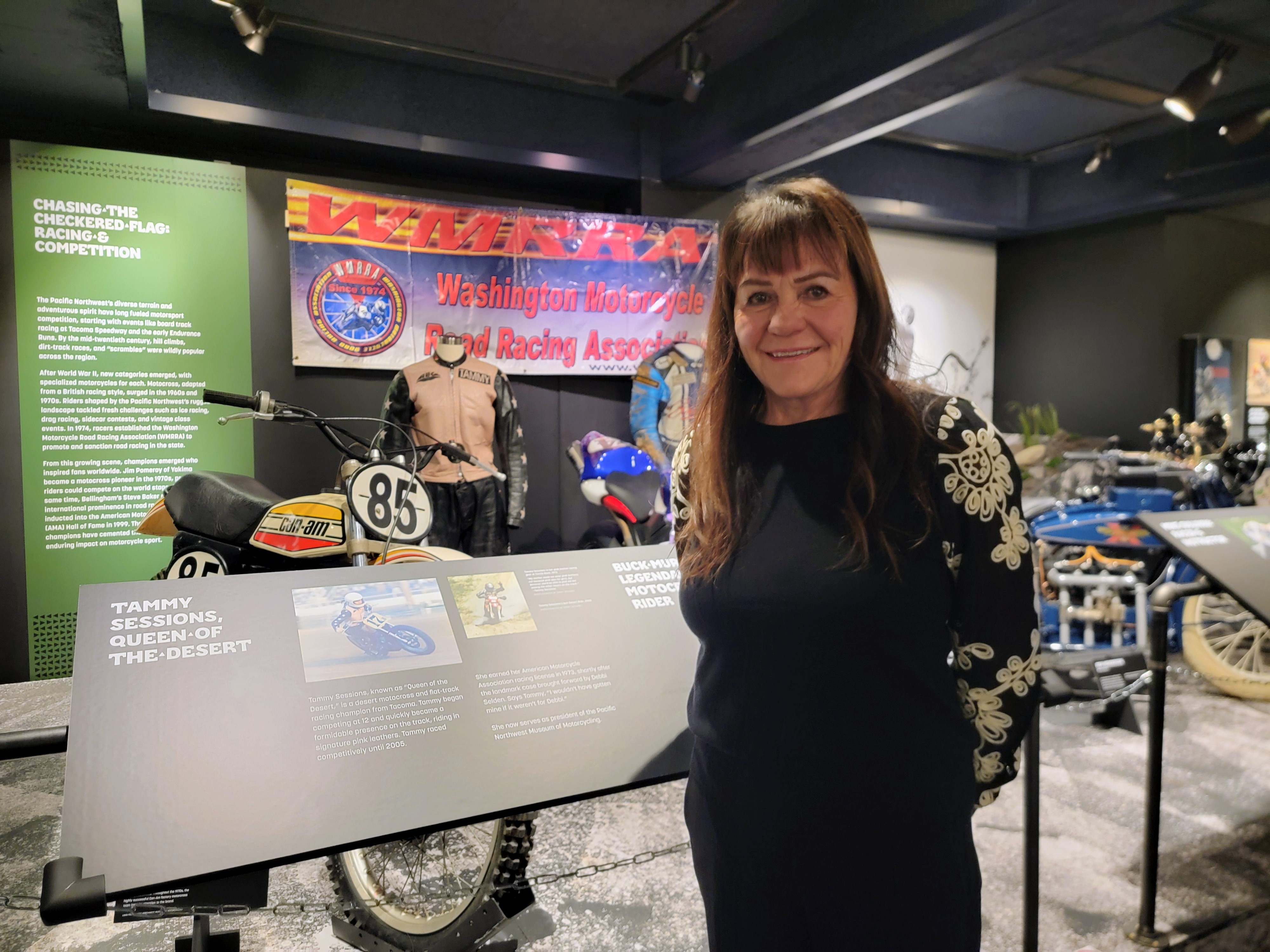 caption: Tammy Sessions stands in front of one of her old racing bikes and leather suit on display in MOHAI on Wednesday, Dec. 3, 2025, as part of the "Kickstands Up!" exhibit celebrating motorcycle culture and history in the Pacific Northwest.