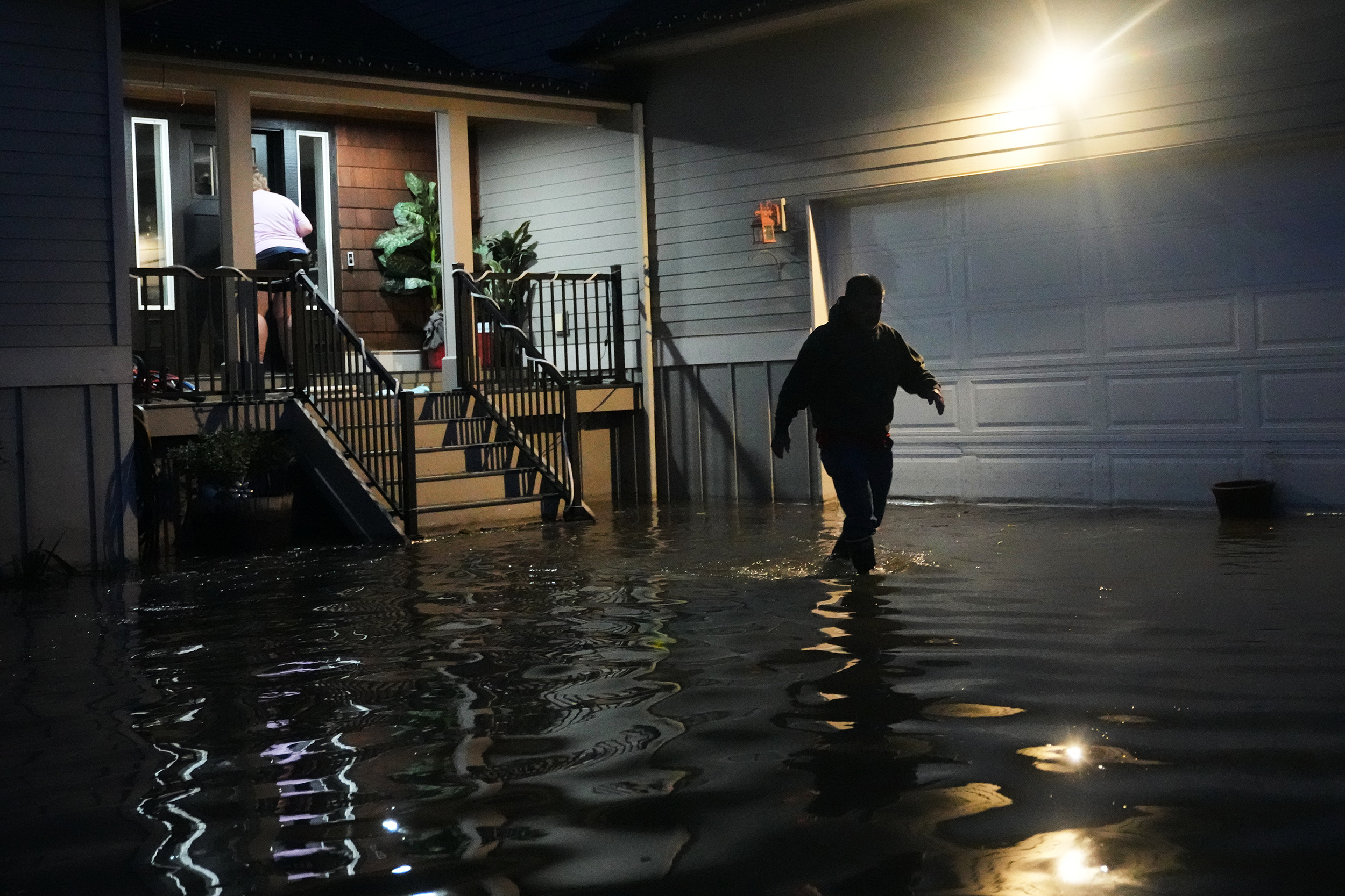 caption: Haji Higa, right, and Lydia Heglin, left, walk through floodwaters at their front door after heavy rains led to historic flooding in the region Saturday, Dec. 13, 2025, in Burlington, Wash. 