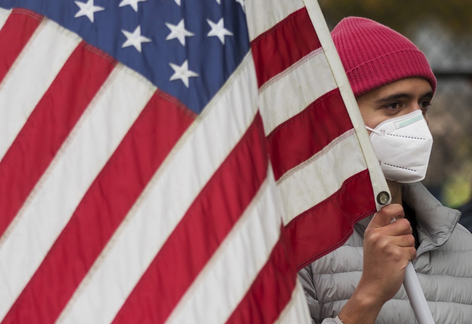 caption: Randolph Lopez holds an American flag while celebrating as a parade of cars drive by and honk their horns after Joe Biden was officially named the president elect on Saturday, November 7, 2020, at the intersection of 10th Avenue and East Pine Street in Seattle.