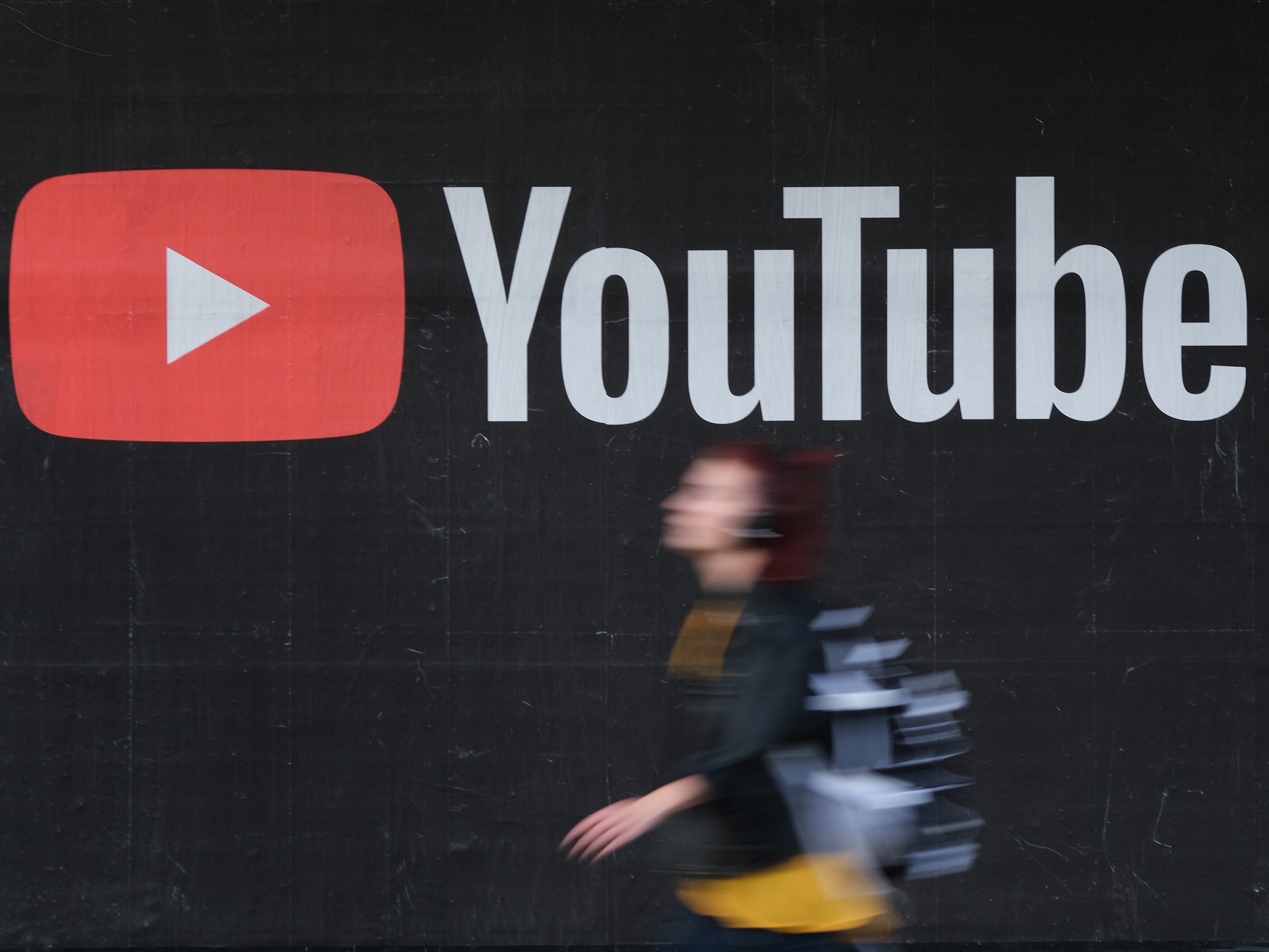 caption: A young woman wearing headphones walks past a billboard advertisement for YouTube in 2019 in Berlin, Germany.