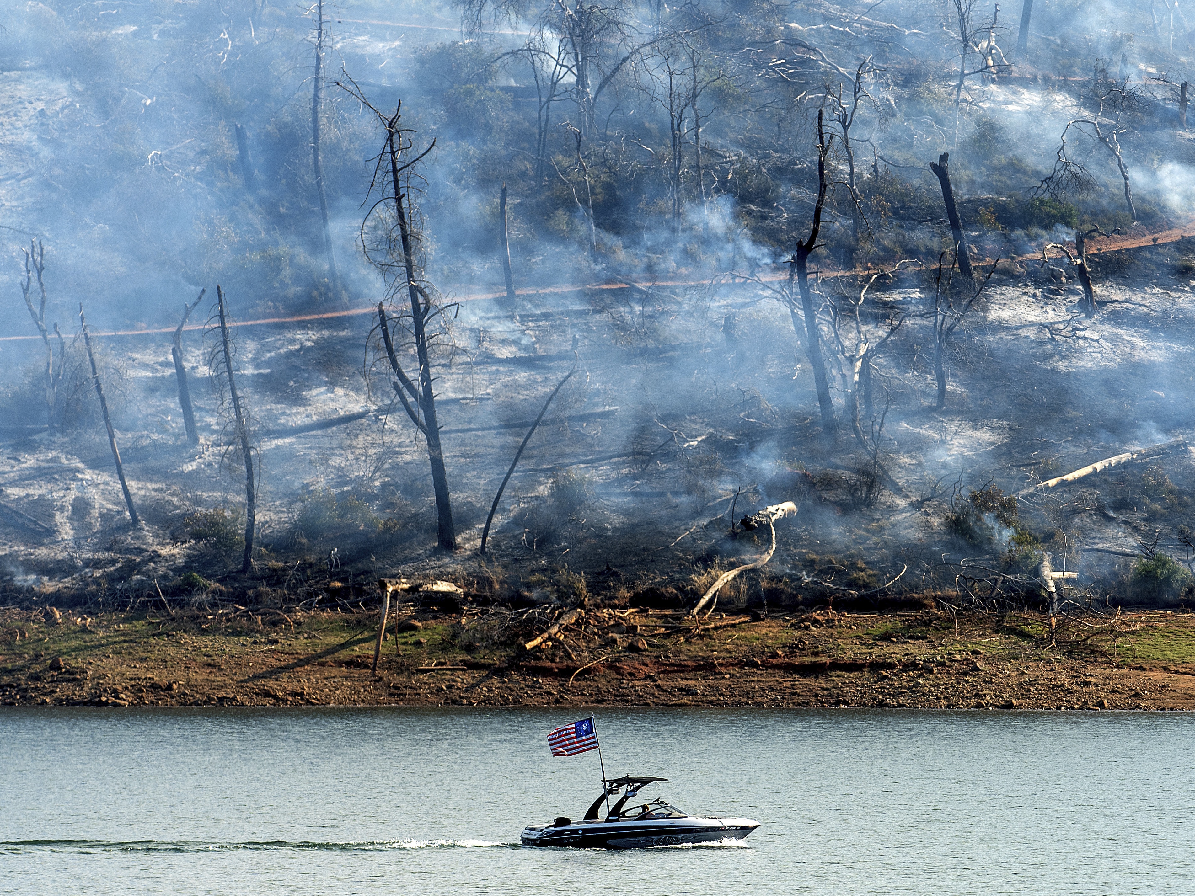 caption: A boat crosses Lake Oroville with a smoldering hillside behind as the Thompson Fire burns in Oroville, Calif., on Wednesday. An extended heatwave blanketing Northern California has resulted in red flag fire warnings and power shutoffs.