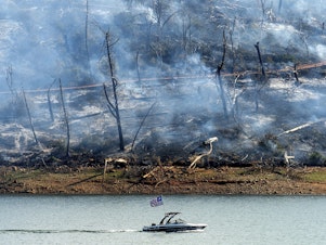 caption: A boat crosses Lake Oroville with a smoldering hillside behind as the Thompson Fire burns in Oroville, Calif., on Wednesday. An extended heatwave blanketing Northern California has resulted in red flag fire warnings and power shutoffs.
