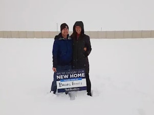 caption: Ari and TR Brooks stood on the land where their new home would be built the day they agreed to buy it back in February of 2021. But the home is still not completed and mortgage rates have risen dramatically.
