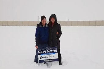 caption: Ari and TR Brooks stood on the land where their new home would be built the day they agreed to buy it back in February of 2021. But the home is still not completed and mortgage rates have risen dramatically.