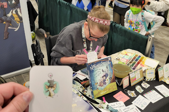 caption: Katie Cook paints custom mini watercolors for fans at Emerald City Comic Con 2024 in Seattle. 