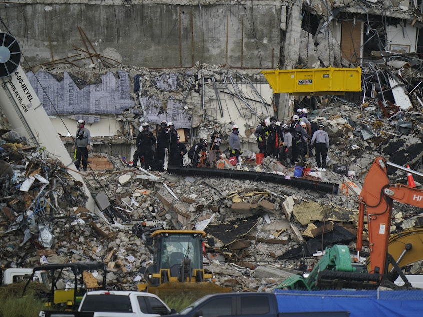 caption: Search and rescue personnel work alongside heavy machinery Wednesday to sift through the rubble at the Champlain Towers South condo building, where scores of people remain missing almost a week after it partially collapsed.