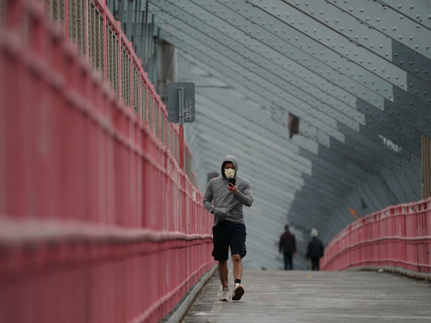 caption: A pedestrian in a face mask crosses the Williamsburg Bridge in New York City last month. U.S. health authorities have announced they're changing the official recommendations on face masks, now urging people to wear them in public spaces to help slow the spread of the coronavirus.