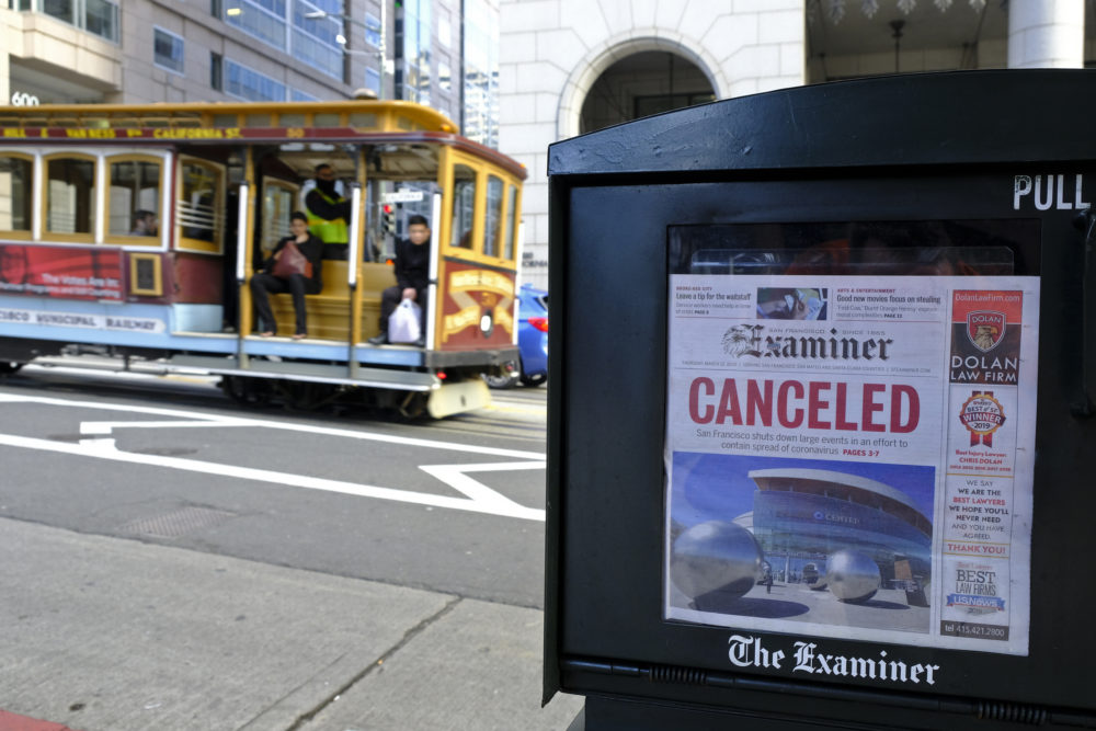 caption: A newspaper headline announcing the closure of large events is displayed as a cable car goes down California Street, Friday, March 13, 2020, in San Francisco. (Eric Risberg/AP Photo)