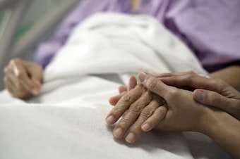 A daughter holds her mother's hand and encourages her while sitting on a hospital bed.