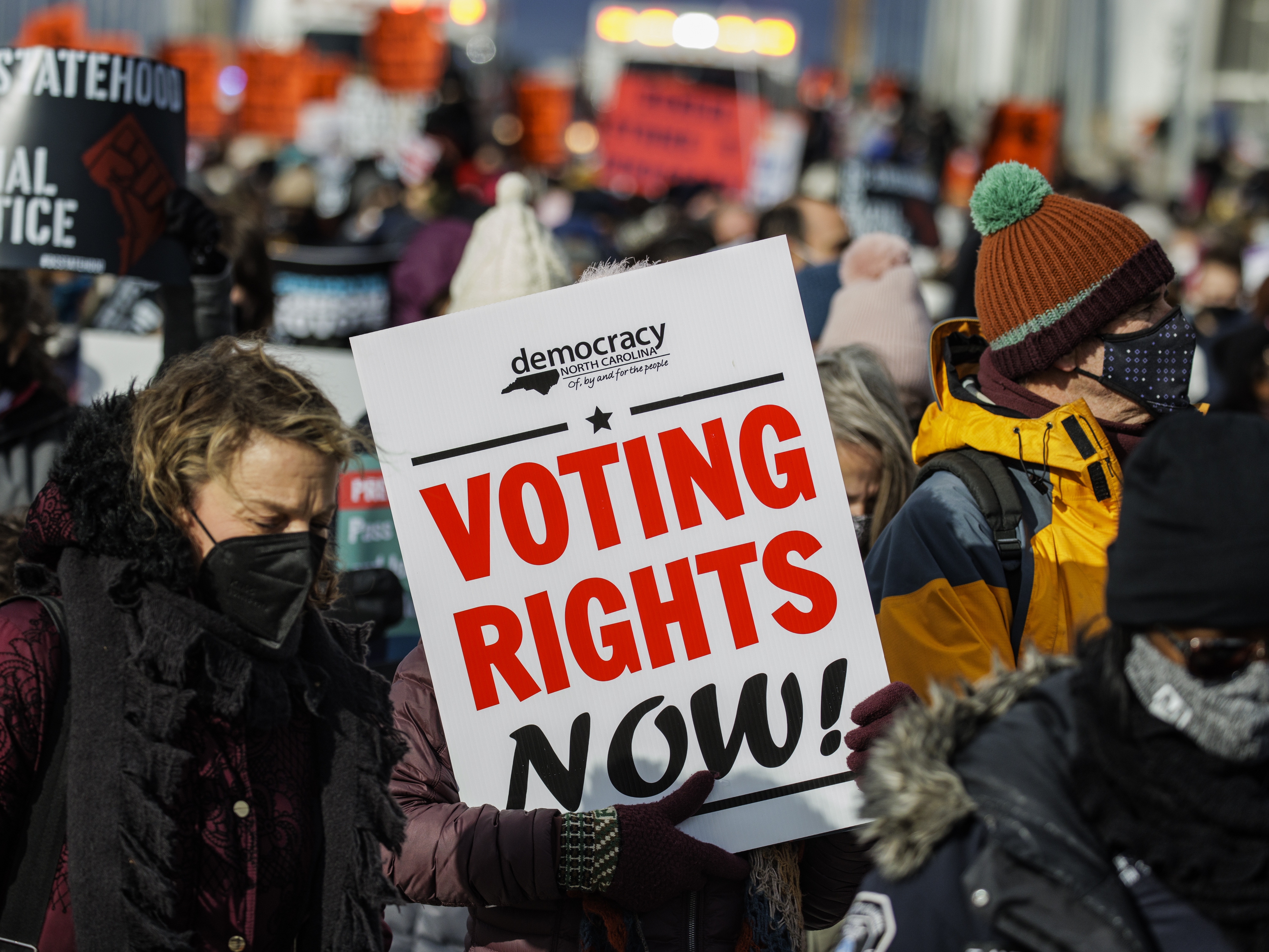 caption: A demonstrator carrying a sign that says "VOTING RIGHTS NOW" walks across the Frederick Douglass Memorial Bridge in 2022 in Washington, D.C.