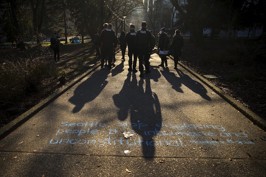 caption: Seattle police officers walk over chalk writing that reads 'Seattle, stop displacing people. It is inhumane and unconstitutional. Martin v. Boise,' before sweeping unhoused people from Denny Park on Wednesday, March 3, 2021, in Seattle. 