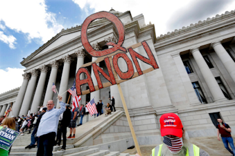 caption: A demonstrator holds a "Q Anon" sign as he walks at a protest opposing Washington state's stay-home order to slow the coronavirus outbreak Sunday, April 19, 2020, in Olympia, Wash. Washington Gov. Jay Inslee has blasted President Donald Trump's calls to "liberate" parts of the country from stay-at-home and other orders that are designed to combat the spread of the coronavirus. Inslee says that Trump is fomenting a potentially deadly "insubordination" among his followers before the pandemic is contained. 