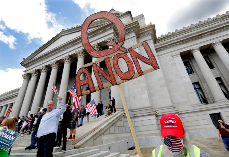 caption: A demonstrator holds a "Q Anon" sign as he walks at a protest opposing Washington state's stay-home order to slow the coronavirus outbreak Sunday, April 19, 2020, in Olympia, Wash. Washington Gov. Jay Inslee has blasted President Donald Trump's calls to "liberate" parts of the country from stay-at-home and other orders that are designed to combat the spread of the coronavirus. Inslee says that Trump is fomenting a potentially deadly "insubordination" among his followers before the pandemic is contained. 