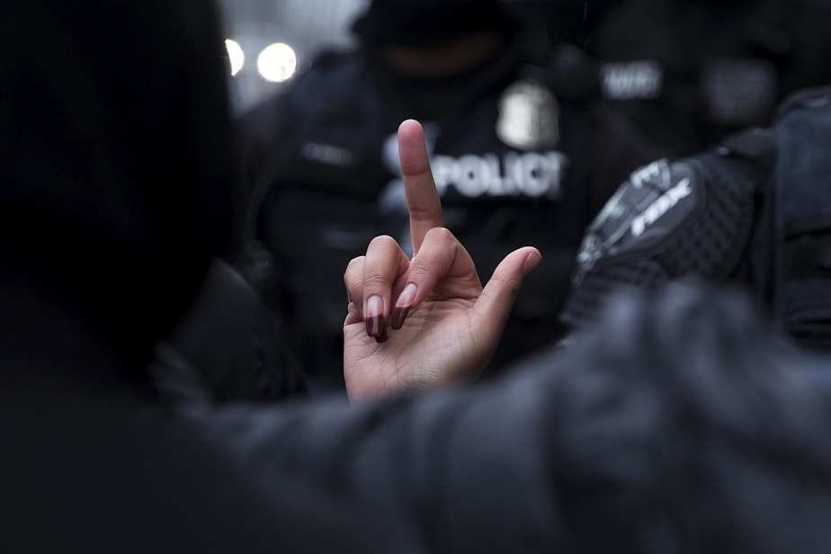 caption: A protester gives Seattle police officers the middle finger after unhoused community members were swept from Cal Anderson Park on Friday, December 18, 2020, in Seattle.  The sweep of the encampment came amid the worst of the ongoing pandemic, with cases of Covid-19 and hospitalizations at their highest levels since the virus first arrived locally. 