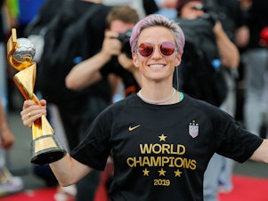 caption: The U.S. Women's National Team is being honored by a ticker tape parade in New York Wednesday. Here, Megan Rapinoe smiles as she holds the FIFA Women's World Cup trophy as the team arrives at the Newark International Airport.