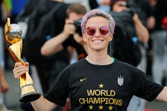 caption: The U.S. Women's National Team is being honored by a ticker tape parade in New York Wednesday. Here, Megan Rapinoe smiles as she holds the FIFA Women's World Cup trophy as the team arrives at the Newark International Airport.