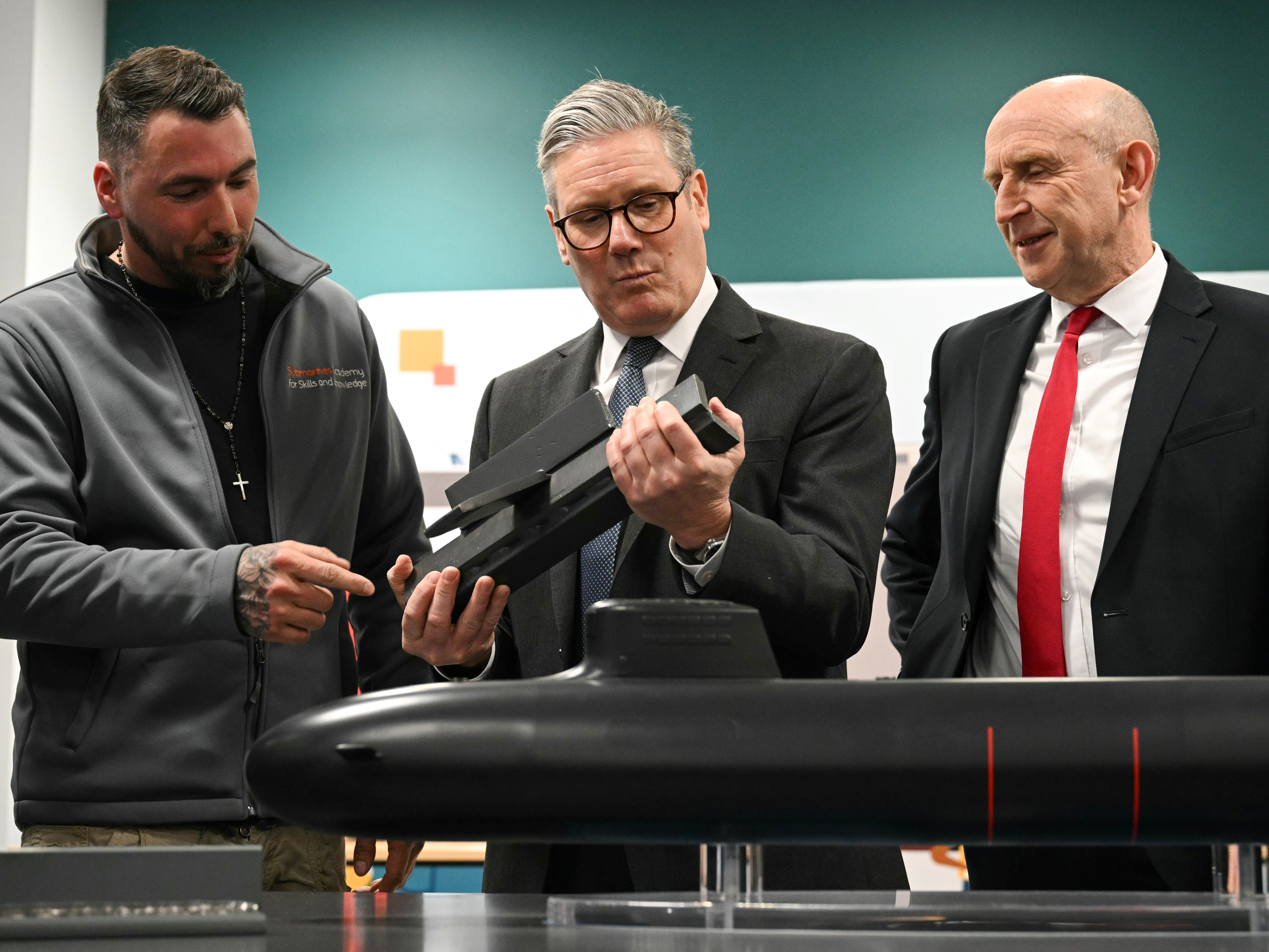 caption: British Prime Minister Keir Starmer (center) and Britain's Defence Secretary John Healey (right) meet with BAE system apprentices as they look at a submarine model during a joint visit on March 20 in Barrow-in-Furness, United Kingdom. Starmer says the U.K. must upgrade its military in response to threats from Russia.