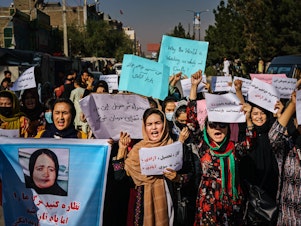 caption: Protesters march in Kabul on Wednesday, a day after the Taliban announced their all-male interim government. At left, a protester carries a sign with a photo showing Banu Negar, a pregnant policewoman who was killed in front of her relatives early this week in Ghor province. Family members accused the Taliban of carrying out the killing.
