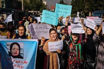 caption: Protesters march in Kabul on Wednesday, a day after the Taliban announced their all-male interim government. At left, a protester carries a sign with a photo showing Banu Negar, a pregnant policewoman who was killed in front of her relatives early this week in Ghor province. Family members accused the Taliban of carrying out the killing.