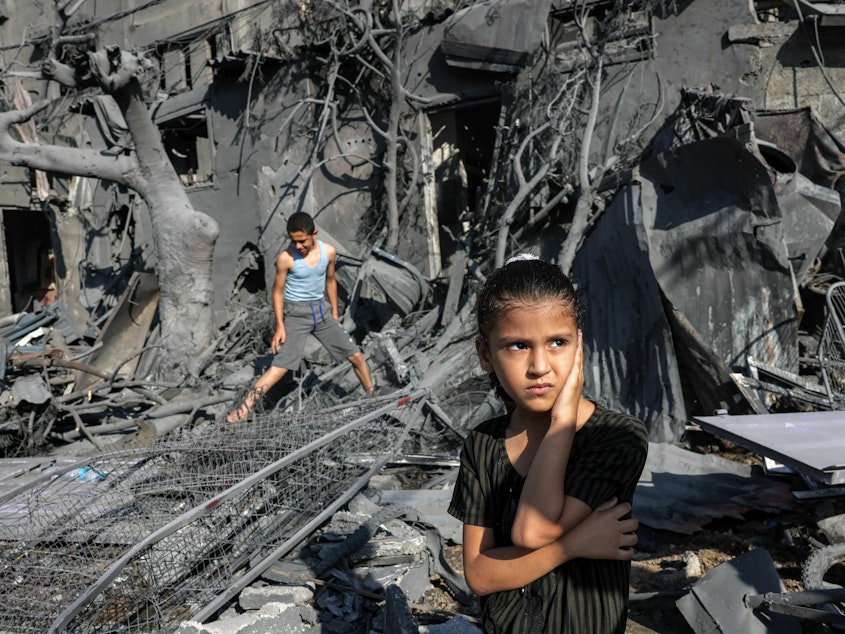 caption: A girl looks on as she stands by the rubble outside a building hit by Israeli bombardment in the southern Gaza Strip on October 31, 2023. Children in Gaza have been exposed to high levels of violence even before the current war, researchers say, increasing their risk of mental health challenges.