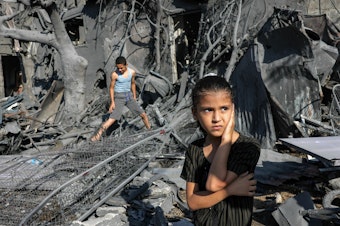 caption: A girl looks on as she stands by the rubble outside a building hit by Israeli bombardment in the southern Gaza Strip on October 31, 2023. Children in Gaza have been exposed to high levels of violence even before the current war, researchers say, increasing their risk of mental health challenges.