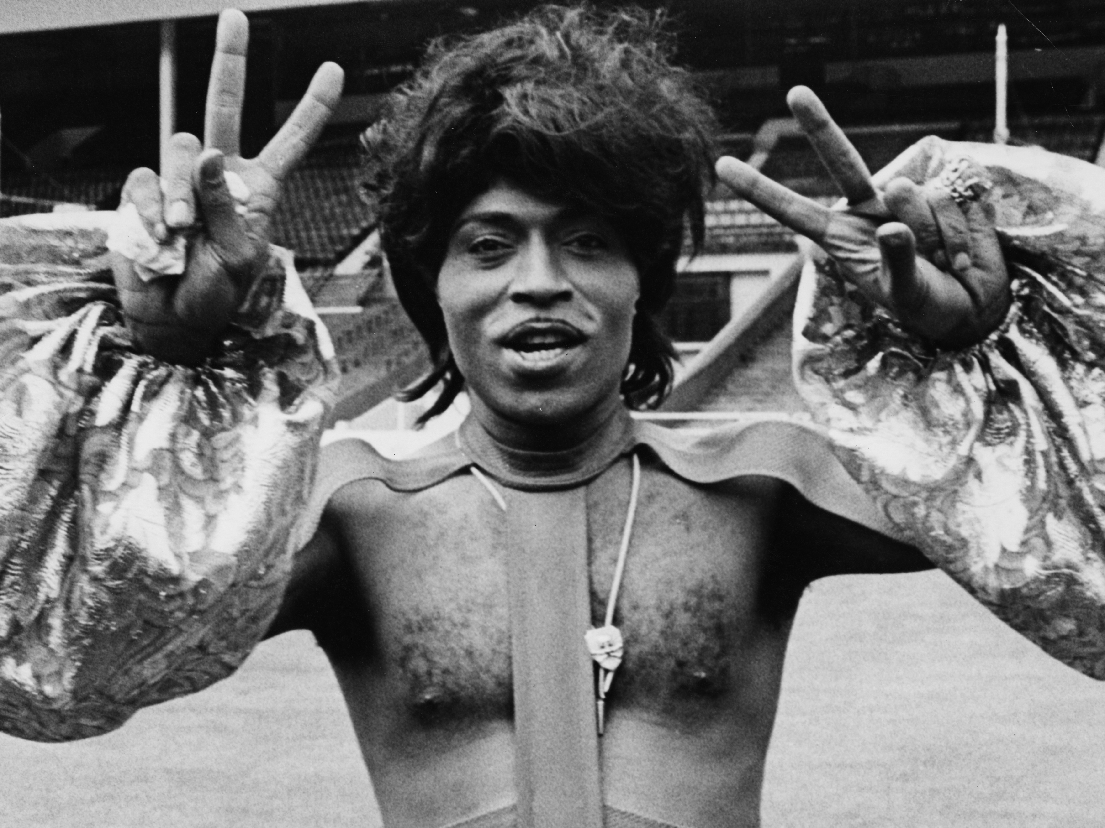 caption: Singer Little Richard making peace sign and wearing an outlandish outfit as he prepares to perform at Wembley Stadium, 1972. (Photo by Rosemary Matthews/Keystone Features/Getty Images)