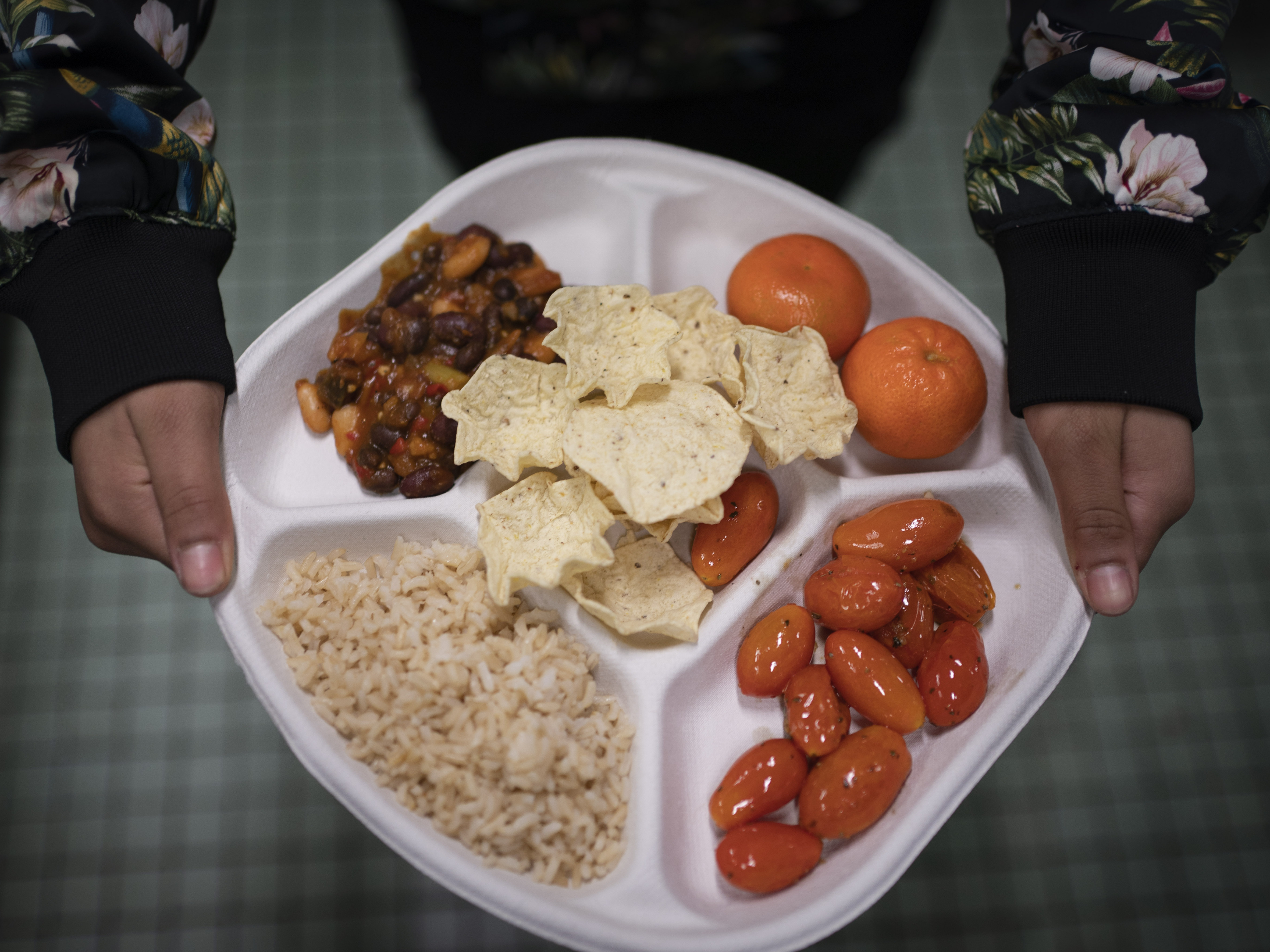 caption: A student carries her plate which consists of three bean chili, rice, mandarins and cherry tomatoes and baked chips during her lunch break at a local public school, Friday, Feb. 10, 2023.