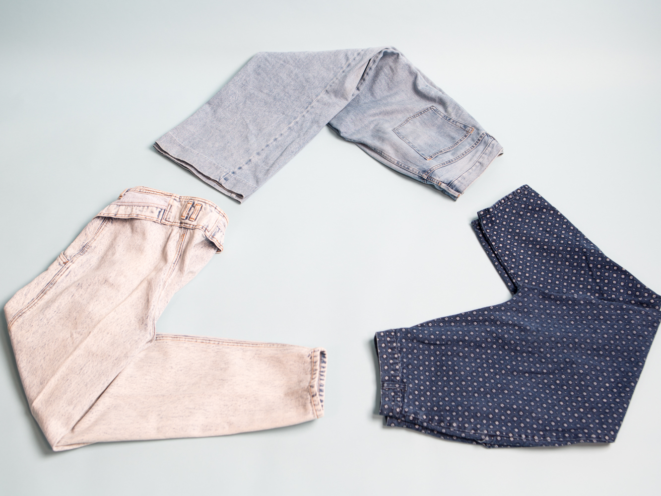 Three pairs of jeans in different shades of denim are arranged in the shape of the "recycle" logo on a light blue backdrop.