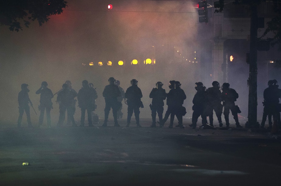 caption: Seattle police officers dressed in riot gear stand in a cloud of tear gas, pepper spray and flash grenade smoke on the fifth day of protests following the violent police killing of George Floyd on Tuesday, June 2, 2020, at the intersection 11th and Pine Street in Seattle.