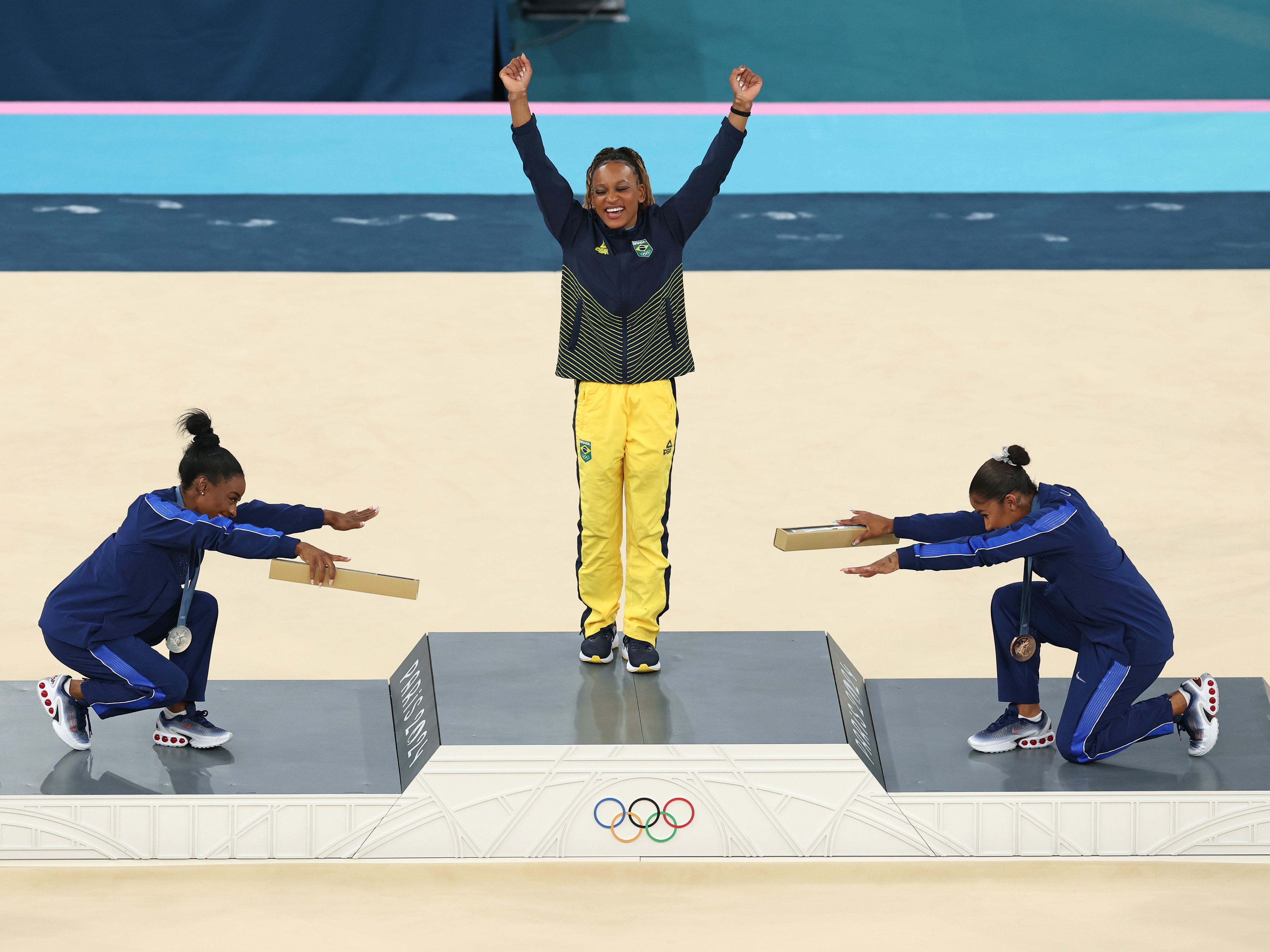 caption: Gymnast Rebeca Andrade (center) of Brazil wins her first gold medal in Paris. Paying homage are silver medalist Simone Biles (left) and bronze medalist Jordan Chiles of the United States. 