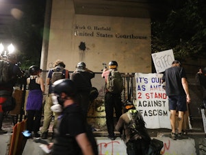 caption: People gather in protest in front of the Mark O. Hatfield federal courthouse in downtown Portland as the city experiences another night of unrest on July 28, 2020 in Portland, Oregon. Protesters in downtown Portland have faced off in often violent clashes with the Portland Police Bureau and, more recently, federal officers.