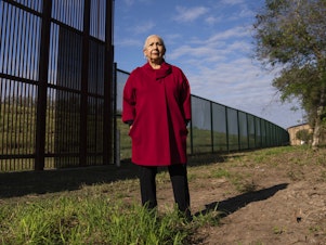 caption: Juliet García, former president of the University of Texas at Brownsville, stands behind the border wall.