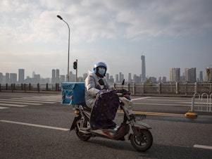 caption: A delivery worker on a motorcycle on an empty street in Wuhan. Despite the city's lockdown measures to prevent the spread of disease, a handful of delivery companies are still in operation. Their workers provide supplies and necessities to residents cooped up in their homes.