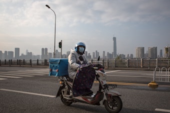 caption: A delivery worker on a motorcycle on an empty street in Wuhan. Despite the city's lockdown measures to prevent the spread of disease, a handful of delivery companies are still in operation. Their workers provide supplies and necessities to residents cooped up in their homes.
