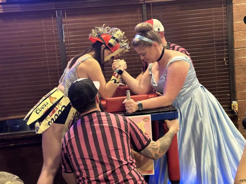 caption: Two women face off at the Arm Wrestling Throwdown, a fundraising event for the Arizona chapter of the Pink Boots Society. The nonprofit raises is a networking group and raises funds to help women succeed in the beer and fermented alcoholic beverage industries. (Jill Ryan/KJZZ)