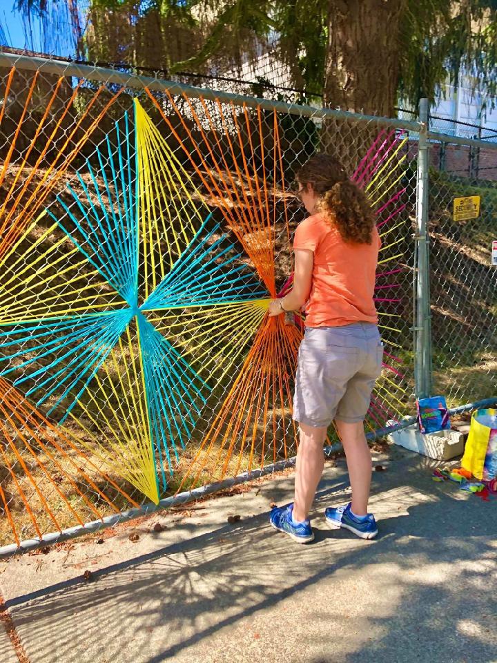 caption: Christy installs one of her brightly colored 'yarnbombs' on a chainlink fence near her home. 