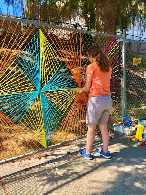 caption: Christy installs one of her brightly colored 'yarnbombs' on a chainlink fence near her home. 