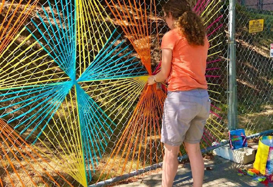 caption: Christy installs one of her brightly colored 'yarnbombs' on a chainlink fence near her home. 