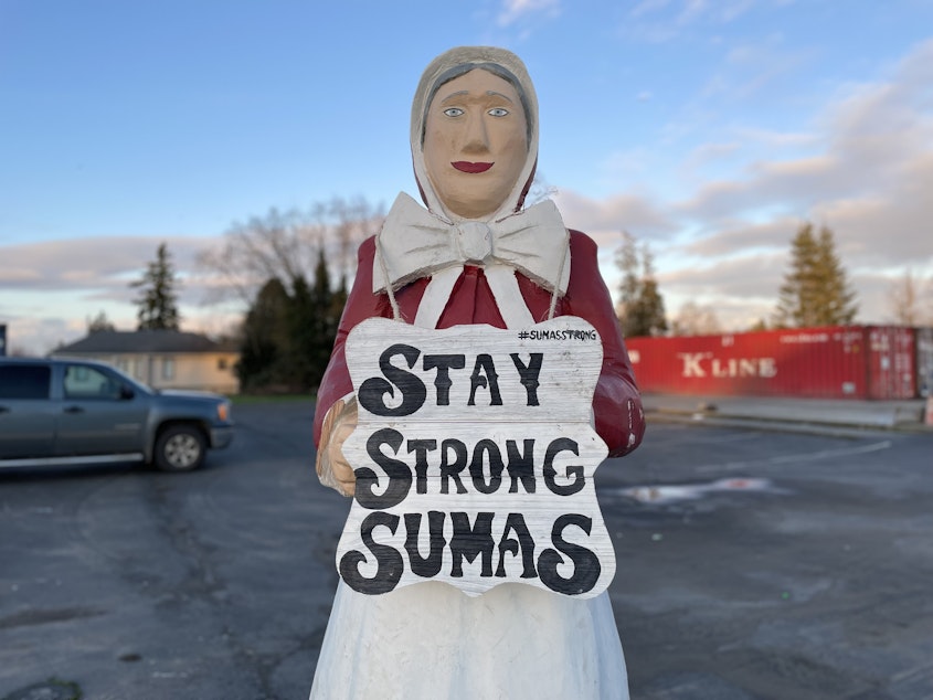 caption: A statue holding a "STAY STRONG SUMAS" sign greets people as they enter the flood-ravaged town. The Nooksack River flooded the town in November 2021. 