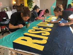 caption: Volunteers paint a banner for the front of the demonstration. Organizers hosted the event to bring supporters together ahead of this year's Women's March, scheduled for Saturday.