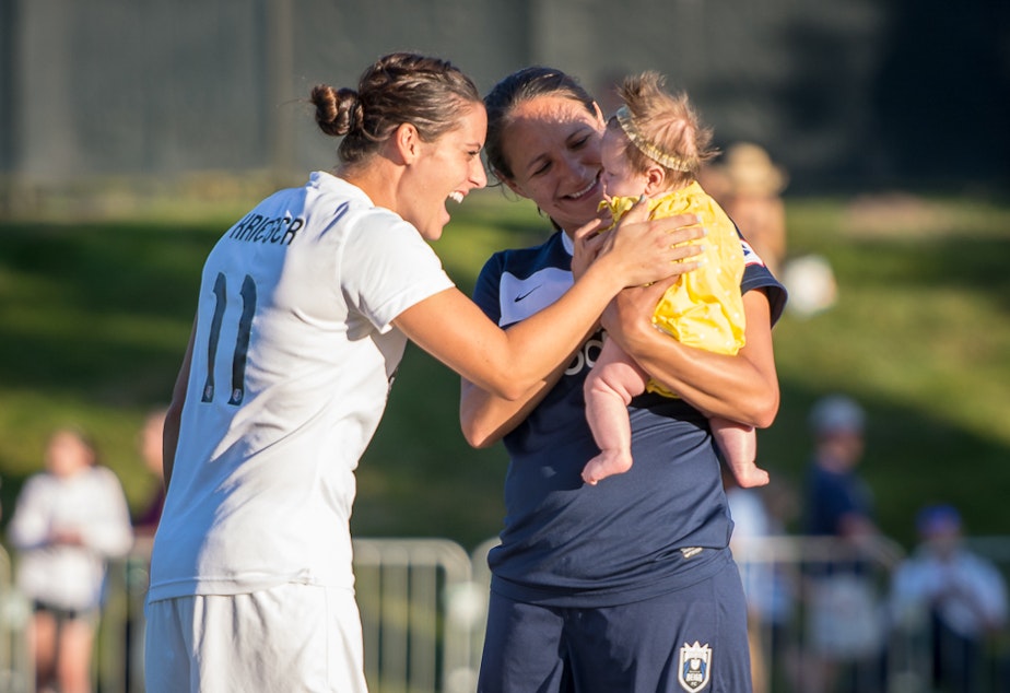 caption: Seattle Reign FC defender Stephanie Cox and her daughter Kaylee are greeted by Washington Spirit opponent (but US National teammate) Ali Krieger at a league game in July. 