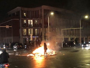 caption: A man walks in front of a fire in a street of The Hague during a demonstration against the Dutch government's coronavirus measures on November 20, 2021.