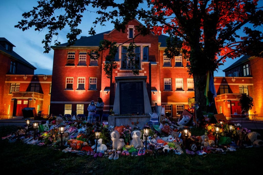 caption: A makeshift memorial to honor the 215 children whose remains have been discovered buried near the facility is seen as orange light drapes the facade of the former Kamloops Indian Residential School in Kamloops, British Columbia, Canada, on June 2, 2021. (Cole Burston/AFP/Getty Images)
